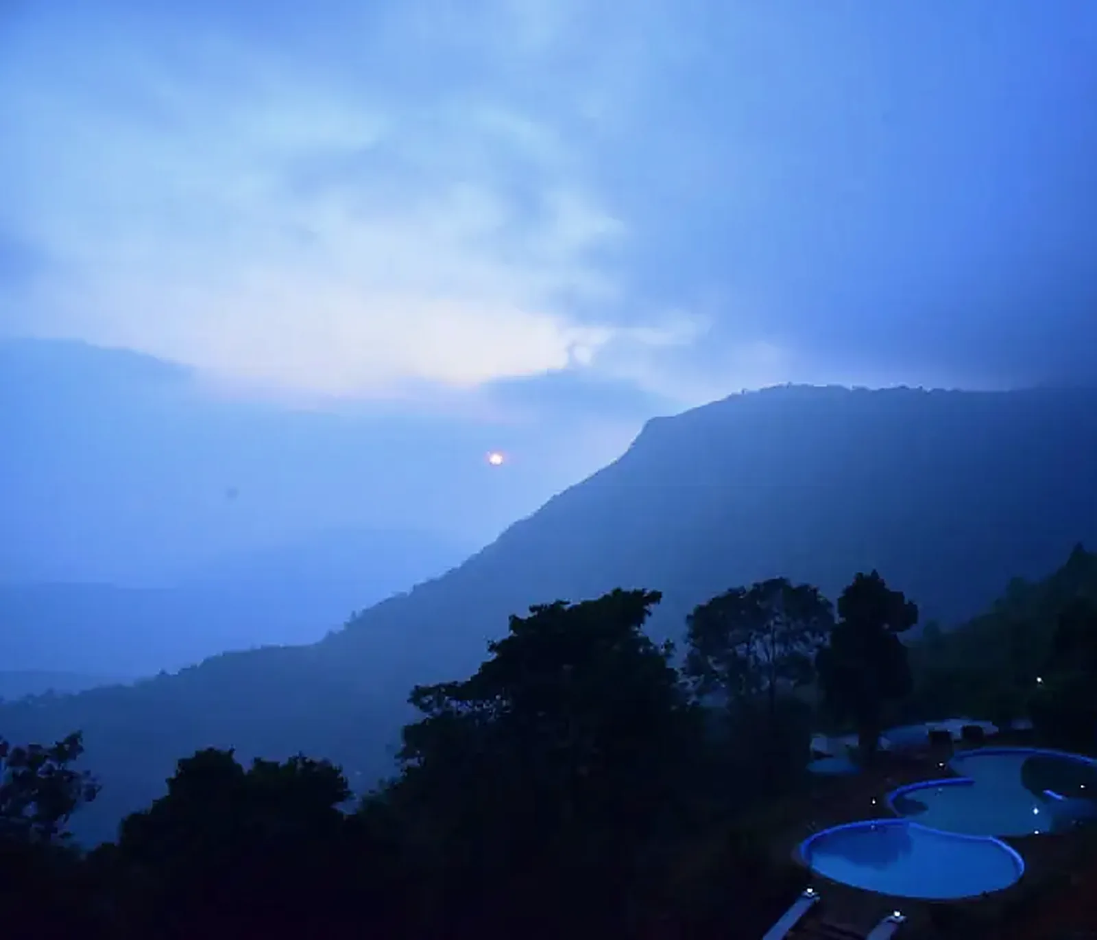 Blue-hour view from the resort: the pool below, forest, and the Paithalmala ridge under cloud